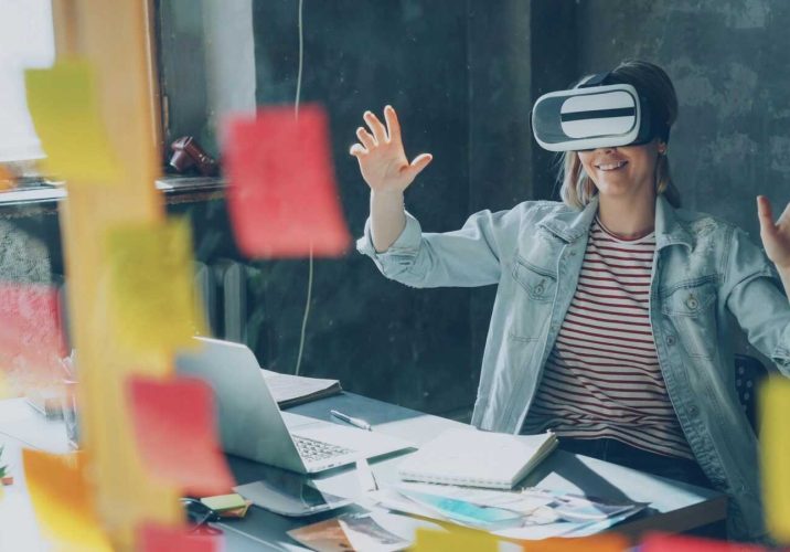 Smiling woman using VR headset in a modern office filled with colorful sticky notes.