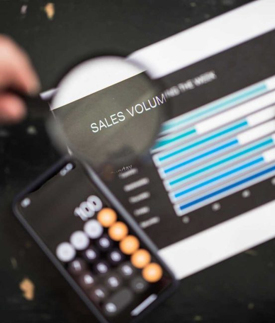 Close-up of a magnifying glass focusing on a sales volume chart next to a smartphone calculator app.